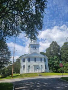 A summertime photo of the Old Meetinghouse, showing a blue sky with puffy white clouds in the background. Lush green foliage surrounds the white Meetinghouse.